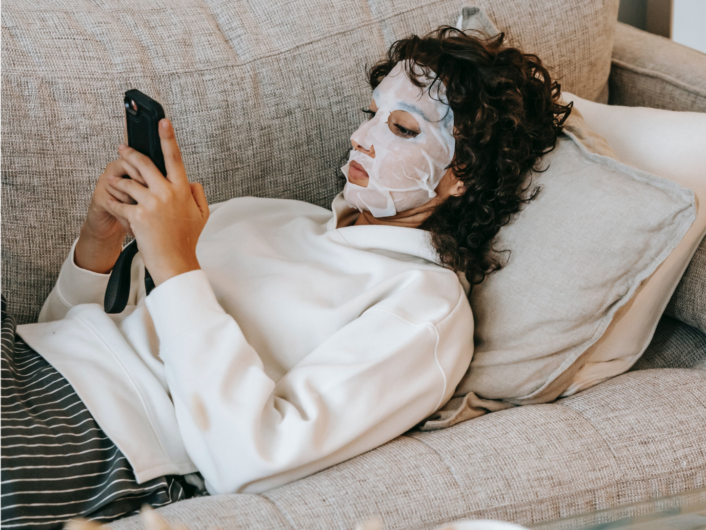 Frau mit Gesichtsmaske liegt entspannt auf der Couch und servt auf dem Handy